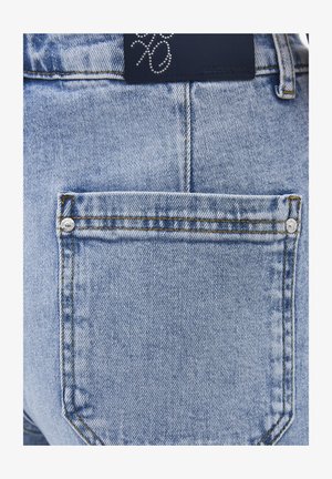 Tasca posteriore di jeans in denim azzurro chiaro con cuciture visibili, due rivetti metallici e una targhetta in pelle nera del marchio con un design di strass sopra.