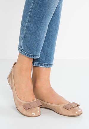 Voeten met glanzende beige ballerina's met stoffen strikjes, gecombineerd met cropped blauwe spijkerbroeken tegen een lichte achtergrond.