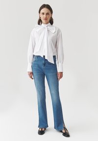 Mujer vistiendo una blusa blanca con un gran lazo en el frente, jeans azules con cortes laterales, zapatos negros con tira en forma de T y pendientes de aro dorados, de pie contra un fondo blanco.
