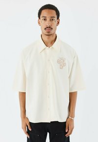 Camisa de manga corta de color crema, hecha de tela suave, con botones en la parte delantera y un bordado de un cactus en el lado izquierdo del pecho.