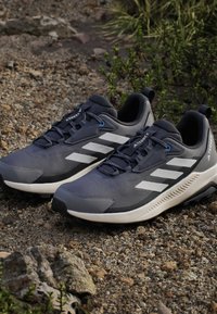 Para czarno-szarych butów biegowych Adidas Terrex z białymi paskami, spoczywających na skalistym terenie na zewnątrz.