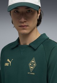 Hanorac verde cu guler, logo auriu și textul "125 JAHRE." Include o șapcă albă cu vizieră verde și un logo Puma.
