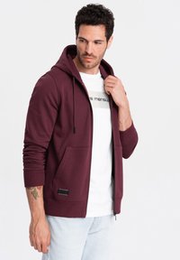 Bordeaux kleurige zip-up hoodie met een kangoeroezak, een capuchon met trekkoord en ribgebreide manchetten. Draagt boven een wit shirt met een bedrukte logo.