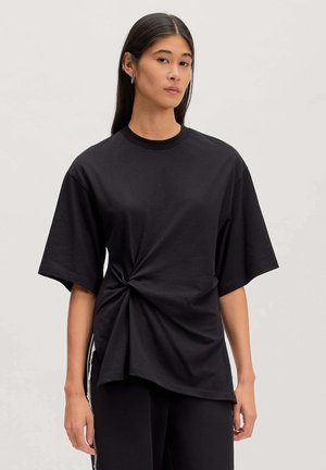 Frau mit langen dunklen Haaren trägt übergroßes schwarzes T-Shirt, seitlich verdreht, und schwarze Hose vor schlichtem hellem Hintergrund.