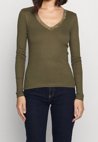 Langärmliges Oberteil in Olivgrün aus strukturiertem Stoff mit V-Ausschnitt und Spitzenbesatz. Kombiniert mit dunkelblauen Jeans; figurbetonte Silhouette.