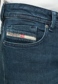 Κοντινή λήψη από την τσέπη μπλε τζιν παντελονιού με ορατή ετικέτα Diesel Industry Denim Division και λεπτομέρειες ραψίματος.