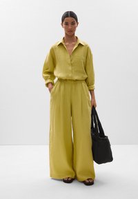 Zweiteiliges Leinen-Outfit in zartem Gelb, bestehend aus einem knöpfbaren Hemd und weiten Hosen mit elastischem Bund; kombiniert mit einer schwarzen Tragetasche.