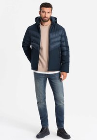 Marineblaue Daunenjacke mit Kapuze, mit diagonal gesteppten Abschnitten, kombiniert mit einem beigefarbenen Sweatshirt, blauen Jeans und schwarzen Stiefeln.