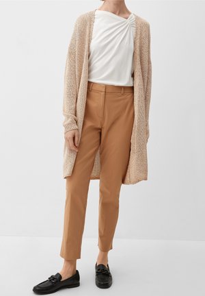 Gilet en maille marron clair sur un haut blanc à encolure froncée, associé à un pantalon beige fuselé et des mocassins noirs.