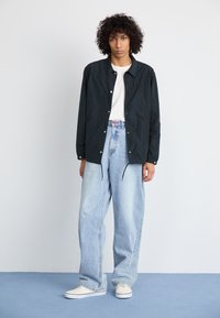 Zwarte lichte jas, wit t-shirt, lichtblauwe wijde jeans en witte sneakers, staand op een blauwe ondergrond tegen een effen achtergrond.