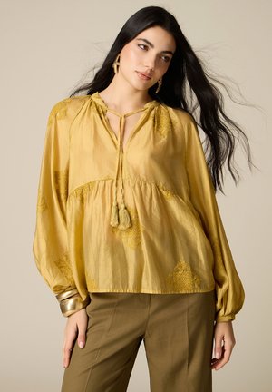 Femme aux longs cheveux noirs portant un chemisier ample jaune moutarde à franges et un pantalon vert olive, accessoirisée avec des bracelets et des boucles d’oreilles dorés.