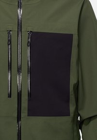 Veste vert olive avec panneau noir, dotée de deux poches zippées et d'une fermeture éclair centrale à l'avant avec une finition texturée. Tissu lisse.
