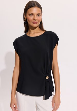 Kobieta ubrana w czarną bluzkę z krótkim rękawem z ozdobnym złotym zapięciem z boku, w połączeniu z białymi spodniami na jednolitym tle.