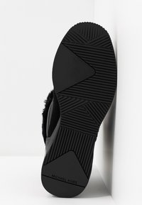 Semelle de chaussure noire avec une adhérence texturée, des motifs géométriques et le marquage "Michael Kors" visible au talon, chaussure posée contre un mur blanc.