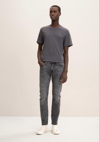 Camiseta gris de cuello redondo, pantalones vaqueros grises desgastados y zapatillas blancas. Modelo de pie, con las manos en los bolsillos, contra un fondo neutro.