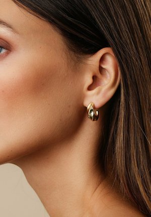 Nahaufnahme des rechten Ohrs einer Frau, die einen verdrehten, glänzenden goldenen Creolenohrring trägt, mit glattem braunem Haar, das hinter dem Ohr steckt.