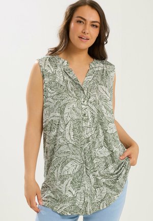 Kvinde iført ærmeløs grøn og hvid bluse med bladmotiv og knapper, poserer med den ene hånd, der holder stof nær hoften.