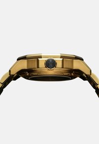 Gouden horloge met een veelzijdige kast, textuur oppervlak en een zwarte kroon met embleem. Het ontwerp is hoekig en robuust.