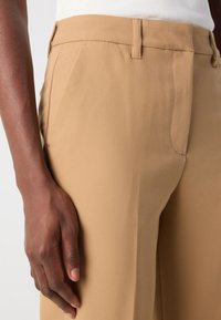 Beige tailored trousers with a smooth, lightweight fabric. Features side pockets and a creased front, displaying a sleek fit and finish.