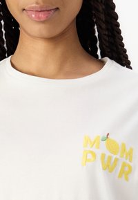 Vit bomulls-t-shirt med gult broderat text "MOM PWR" och en liten citron-grafik. Rund halsringning och mjuk textur.