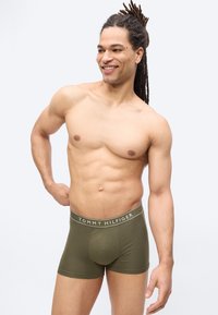 Homme torse nu aux dreadlocks souriant, portant un boxer vert olive Tommy Hilfiger, debout devant un fond clair uni.