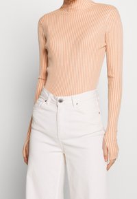 Maglione a maniche lunghe con collo alto in morbido tessuto a costine color pesca, dal taglio slim, abbinato a jeans bianchi a vita alta. Strisce verticali di texture visibili.