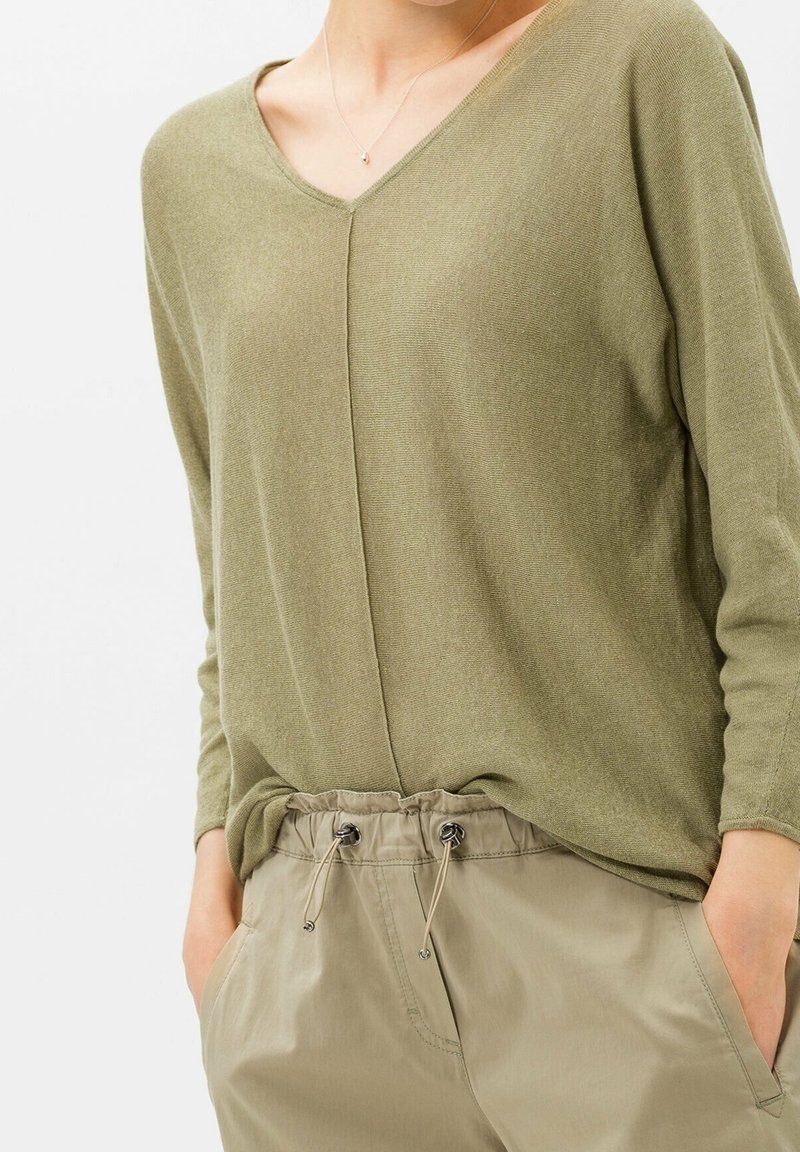 Femme portant un haut à manches longues col en V vert olive rentré dans un pantalon beige à cordon de serrage, les mains dans les poches, montrée du cou aux hanches.