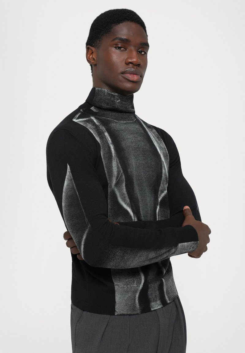 Uomo con le braccia incrociate che indossa un maglione a collo alto nero e grigio con un motivo e pantaloni grigi, davanti a uno sfondo chiaro e semplice.
