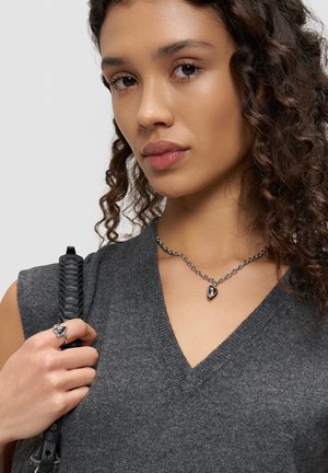 Junge Frau mit lockigem Haar, die einen grauen V-Ausschnitt-Pullover sowie eine silberne Halskette und einen Ring in Herzform trägt und einen schwarzen Riemen hält.