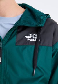 Teal jas met een zwart bovenpaneel en capuchon, voorzien van een ritsluiting en een merklogo. Het materiaal lijkt licht en soepel.