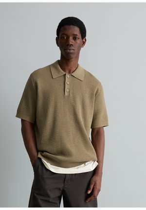 Junger Mann trägt beige strukturiertes kurzärmeliges Polohemd über weißem Unterhemd und dunkler Hose, steht vor grauem Hintergrund.