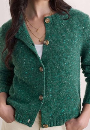 Woman wearing a green speckled buttoned cardigan over a white top, paired with beige pants and layered gold necklaces.