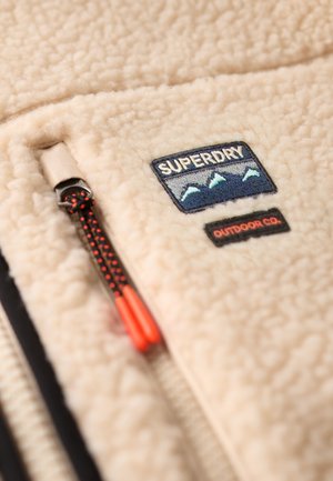 Beige fleecejack met een patch van Superdry Outdoor Co., met berggraphic en een zwarte ritssluiting met rood uiteinde aan het trekkoord.