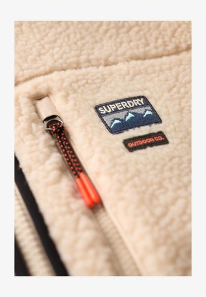 Beige fleecejakke med et Superdry Outdoor Co.-mærke, der har bjerggrafik, og en sort lynlås med røde trækstropper.