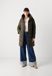 Olijfgroen pufferjack met een capuchon, gecombineerd met wijde blauwe jeans met een rafelrand. Model staat tegen een neutrale achtergrond.