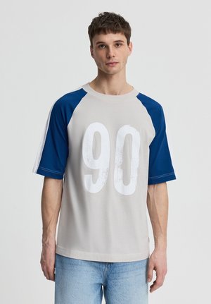 Giovane uomo che indossa una camicia beige con maniche raglan blu e un grande numero bianco "90" sul davanti, abbinata a jeans azzurri.