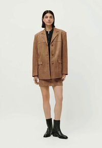 Donna con capelli corti scuri che indossa un blazer oversize in suede marrone, una gonna corta abbinata, un top nero e stivali alla caviglia neri, in piedi di fronte a uno sfondo bianco.