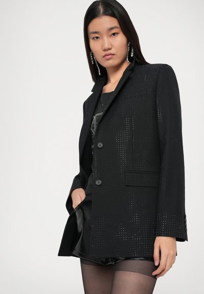 Blazer nero con design strutturato, caratterizzato da dettagli perforati, due bottoni e una tasca. Indossato sopra un top e una gonna neri.
