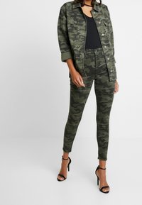 Chaqueta de camuflaje y jeans ajustados en varios tonos de verde, combinados con sandalias de tacón alto negras con tiras. Textura de tela suave.