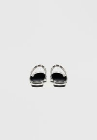 Ασημί σανδάλια με επίπεδη σόλα, με ανοιχτά δάχτυλα και διακοσμητικό λουράκι. Μαύρη υφασμάτινη εσωτερική σόλα με ελάχιστες λεπτομέρειες.