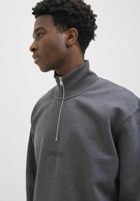 Junger Mann mit kurzen lockigen Haaren, der einen grauen Sweatshirt mit Reißverschluss und einem gerippten hohen Kragen trägt, blickt nach links vor einem schlichten Hintergrund.