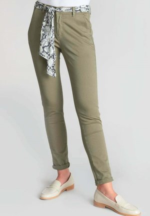 Olivegrüne Slim-Fit-Hosen mit einem Schlangenmotiv-Gürtel. Aus glattem Baumwollstoff, knöchellang, mit aufgerollten Säumen. Begleitet von weißen Slippers.
