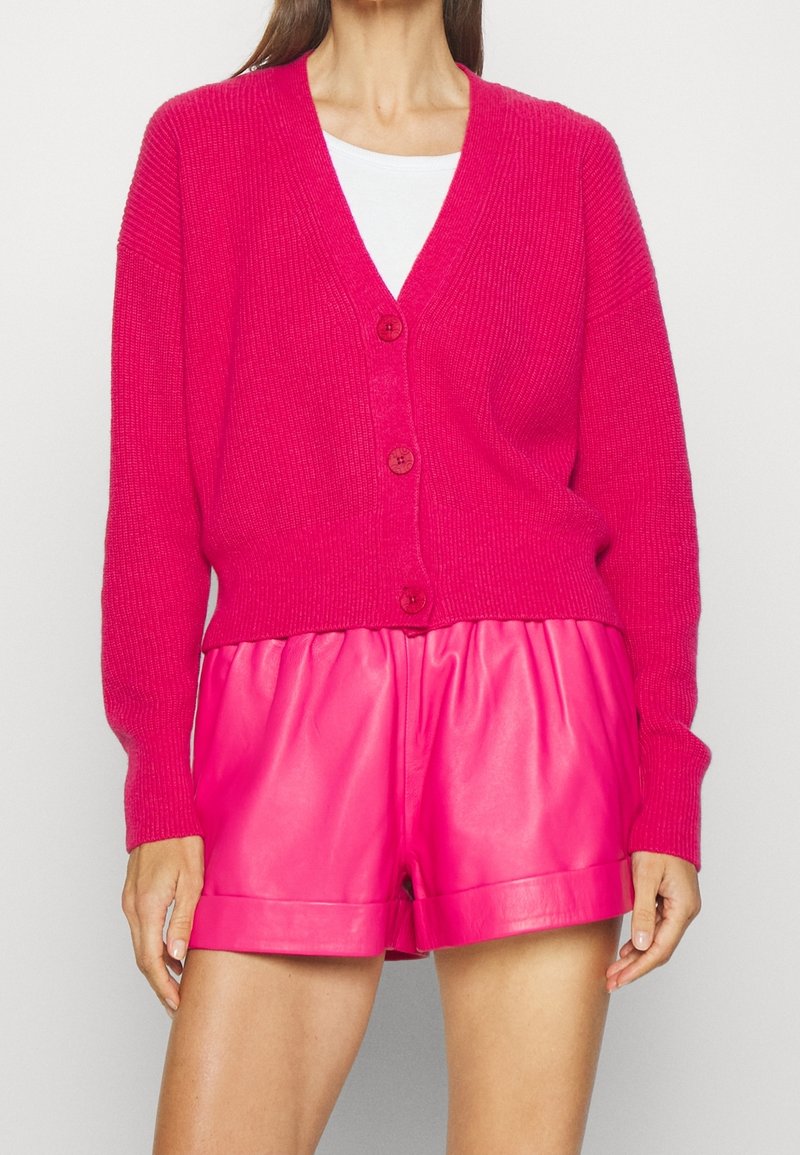 Różowy sweter kardigan z dekoltem w serek i zapięciem na guziki, z prążkowanymi mankietami i dołem. Połączony z dopasowanymi, błyszczącymi skórzanymi szortami.