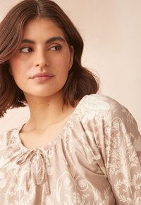 Světlounce hnědá halenka s květinovým bílým potiskem, řaseným výstřihem a stahovacím detailem. Měkká textura a volný střih.