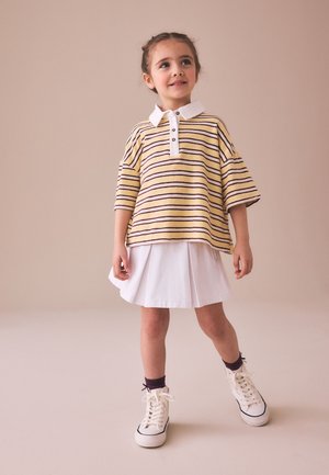 Niña joven de pie mirando hacia arriba, vistiendo una camisa a rayas amarilla con cuello blanco, falda blanca, calcetines oscuros y zapatillas blancas.