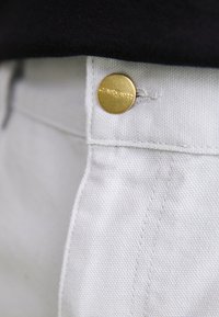 Witte denim broek met een zichtbare gouden knoop met het reliëf van de merknaam, close-up van de tailleband en de textuur van de stof.