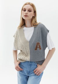 Gilet lavorato a maglia in beige e grigio, con un motivo a treccia. Maniche corte, scollatura a V e una "A" ricamata sul lato grigio.