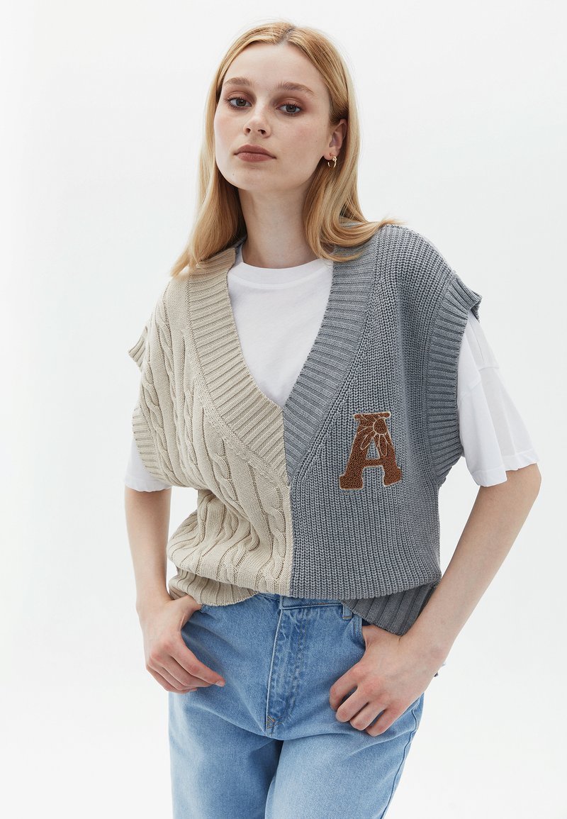 Gilet lavorato a maglia in beige e grigio, con un motivo a treccia. Maniche corte, scollatura a V e una "A" ricamata sul lato grigio.