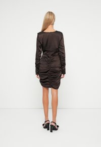 Rochie din satin maro cu mâneci lungi, prezentând detalii pliate la spate și o siluetă ajustată. Sandale cu toc înalt negre vizibile.