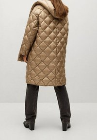 Abrigo acolchado marrón con capucha, que presenta un patrón de diamantes. Llevado sobre jeans oscuros y botas de tacón cuadrado. Textura lisa y brillante.